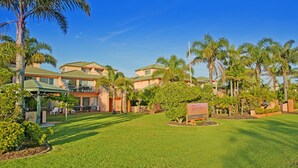 A hotel with a green lawn, palm trees, and a sign indicating a vacancy. - Tuscany Apartments (Merimbula)