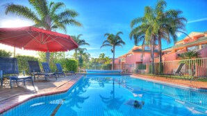 A swimming pool with lounge chairs and umbrellas, surrounded by palm trees and a building with balconies. - Tuscany Apartments (Merimbula)