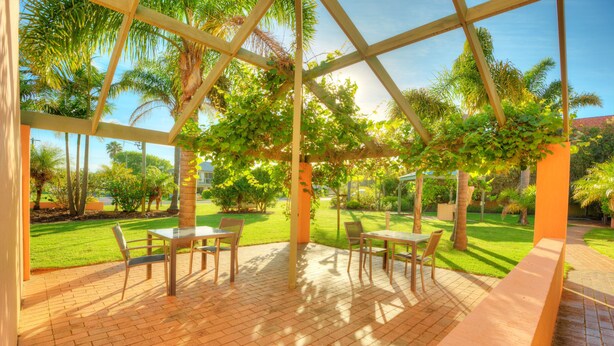 A covered patio with brick flooring, tables, and chairs, surrounded by lush greenery and palm trees. - Tuscany Apartments (Merimbula)