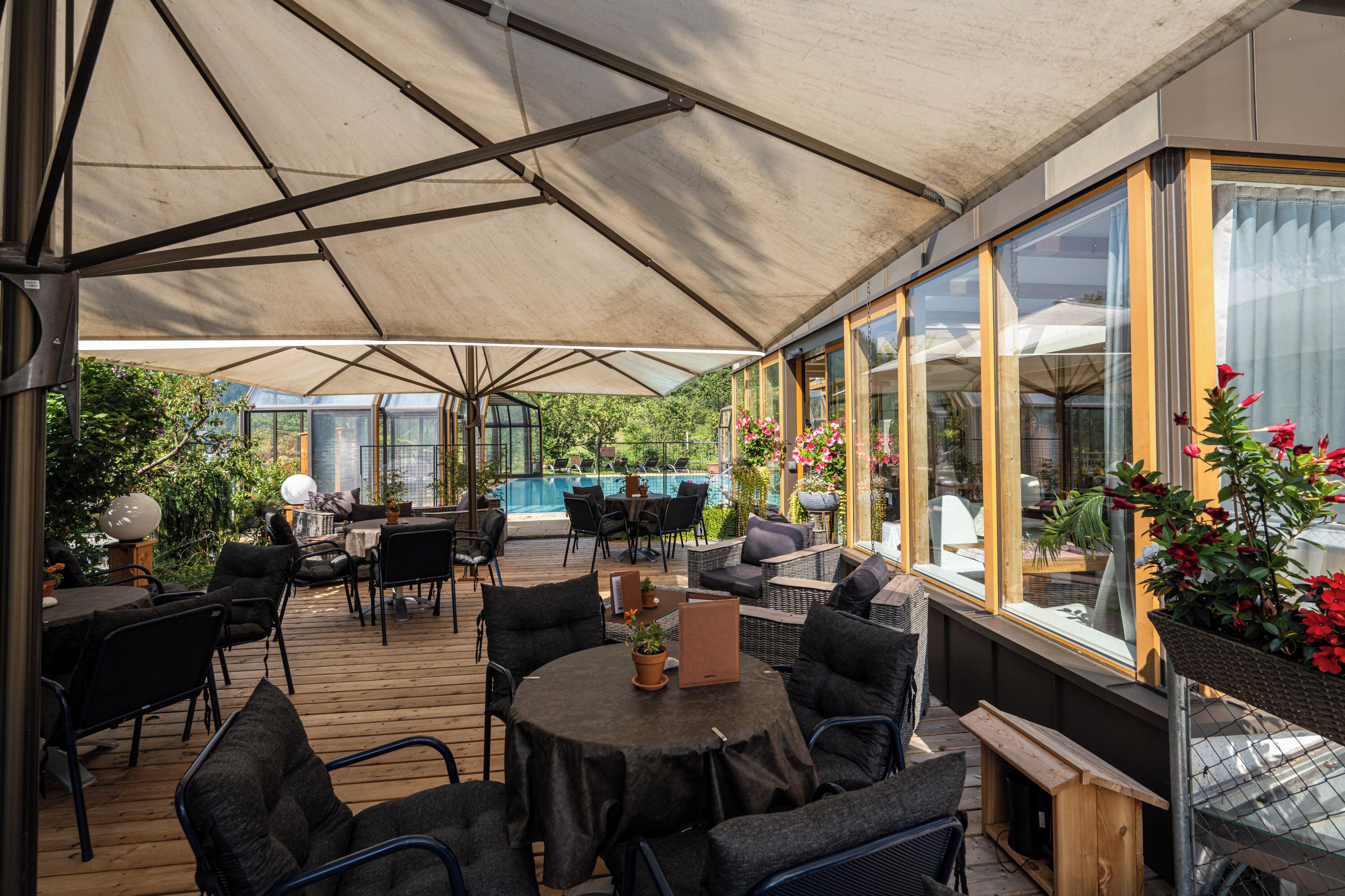 Un espace extérieur aménagé en terrasse, avec des tables et des chaises, une piscine en arrière-plan et de grandes fenêtres.
