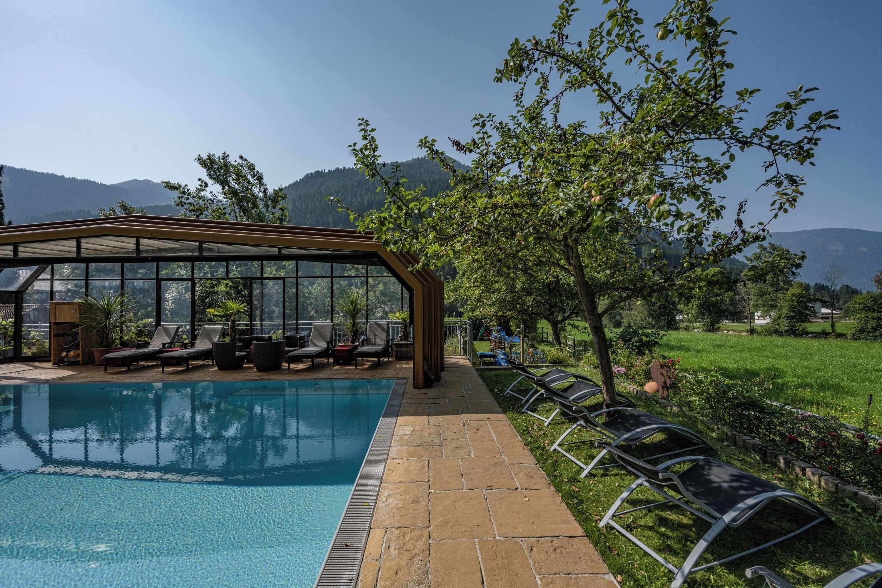 Un área de piscina con sillones de descanso y una zona de estar cubierta, rodeada de follaje y montañas al fondo.