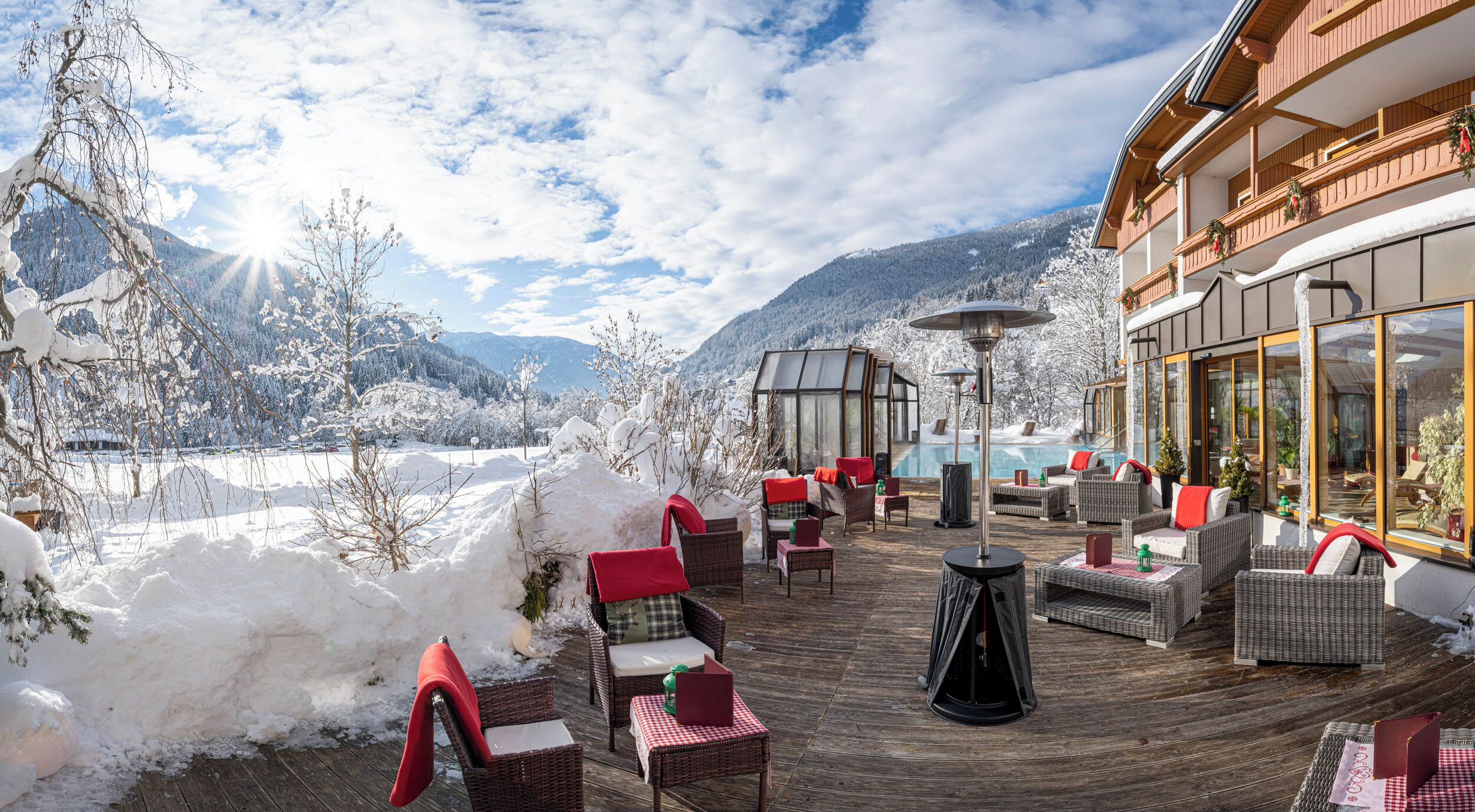 A wooden deck with wicker furniture and a glass-enclosed pool area, overlooking a snowy mountain landscape.