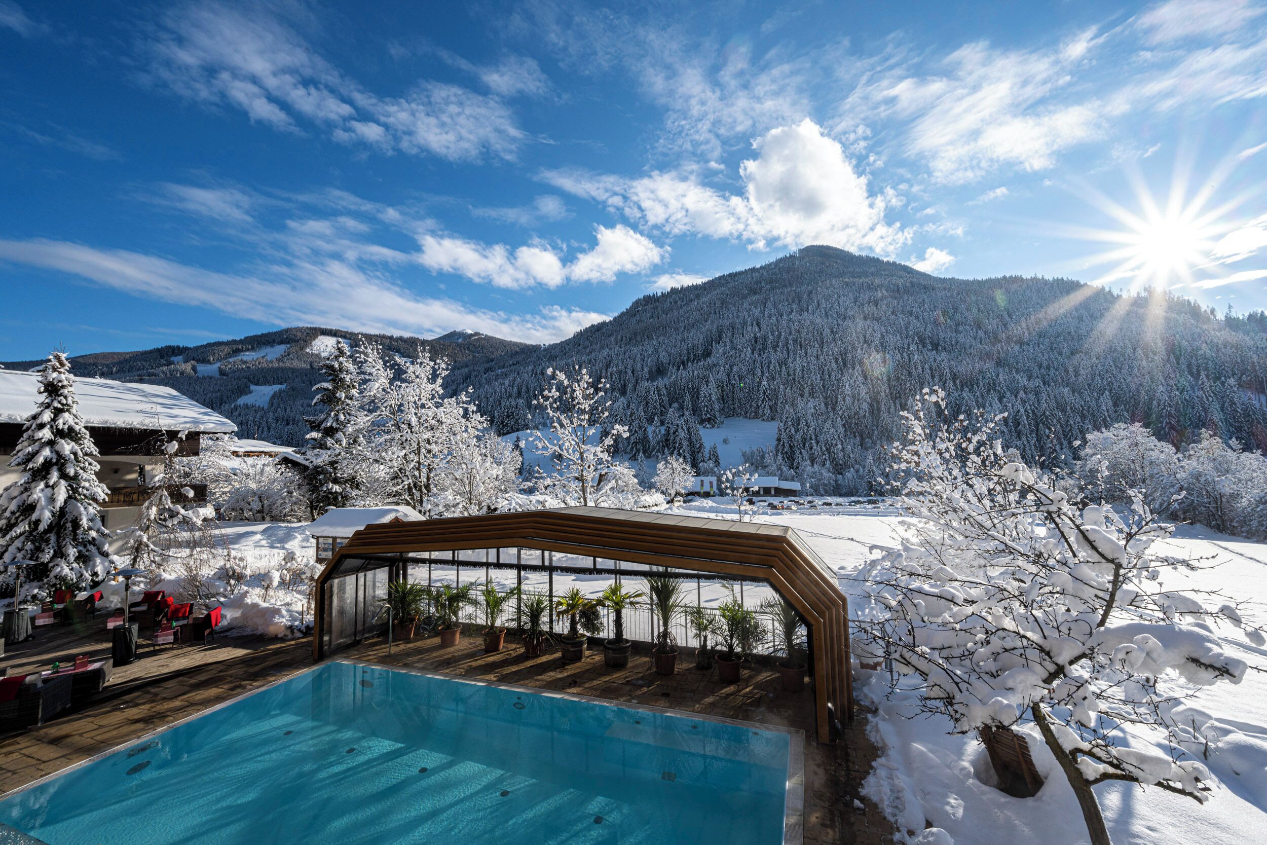 Un área de piscina con una terraza de madera, rodeada de árboles nevados y montañas al fondo.
