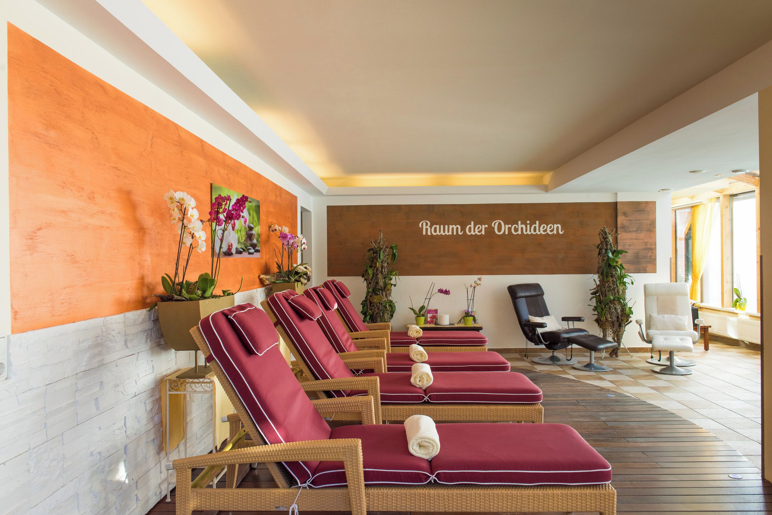 Un cuarto de spa con sillones de mimbre, una pared de madera con el texto 'Raum der Orchideen' y plantas en macetas.