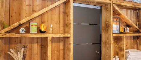 A wooden interior with shelves holding toiletries, a vase with dried flowers, and a framed picture.