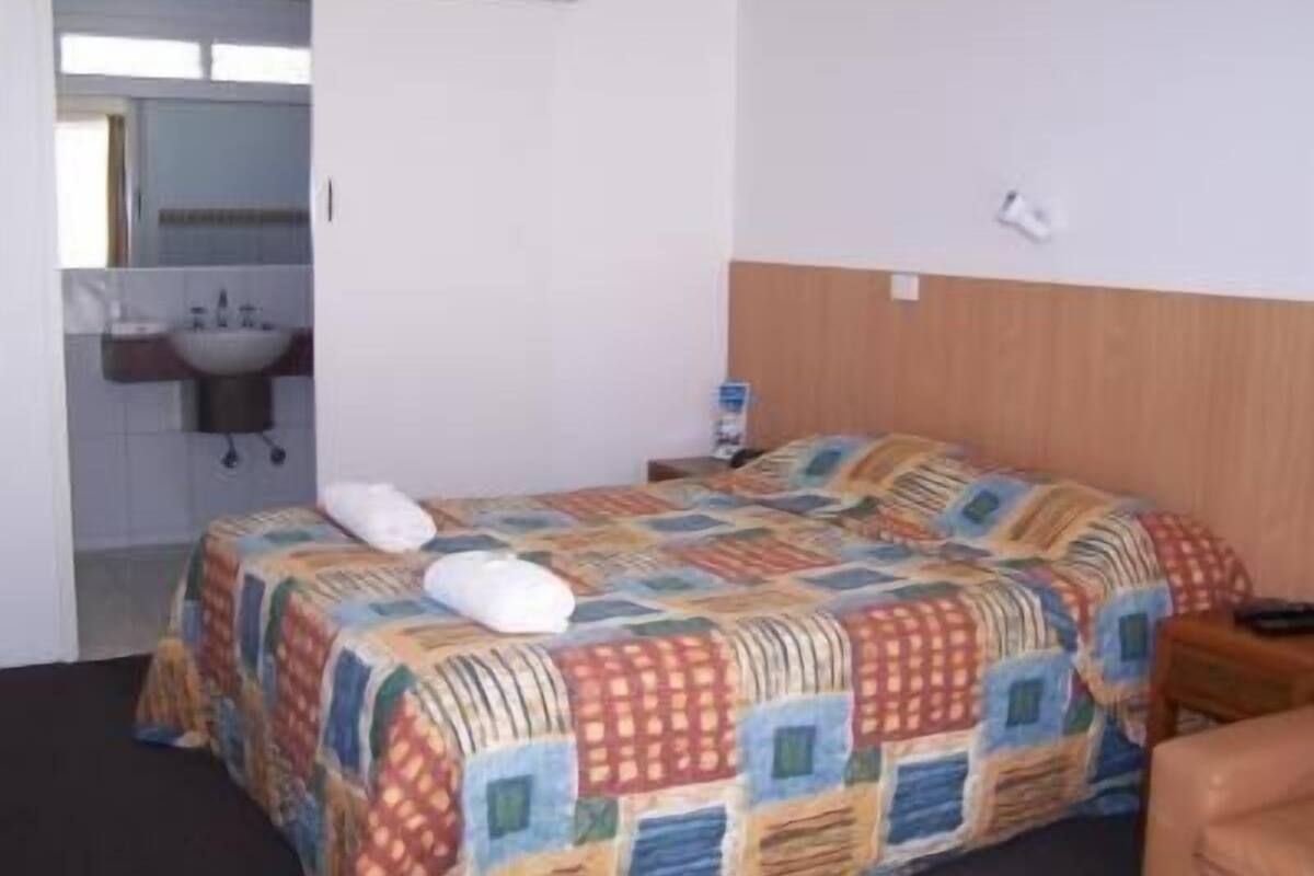A hotel room with a bed, a wooden headboard, a nightstand, a bedside table, a white towel, and a bathroom visible through an open door.