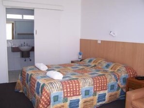 A hotel room with a bed, a wooden headboard, a nightstand, a bedside table, a white towel, and a bathroom visible through an open door.