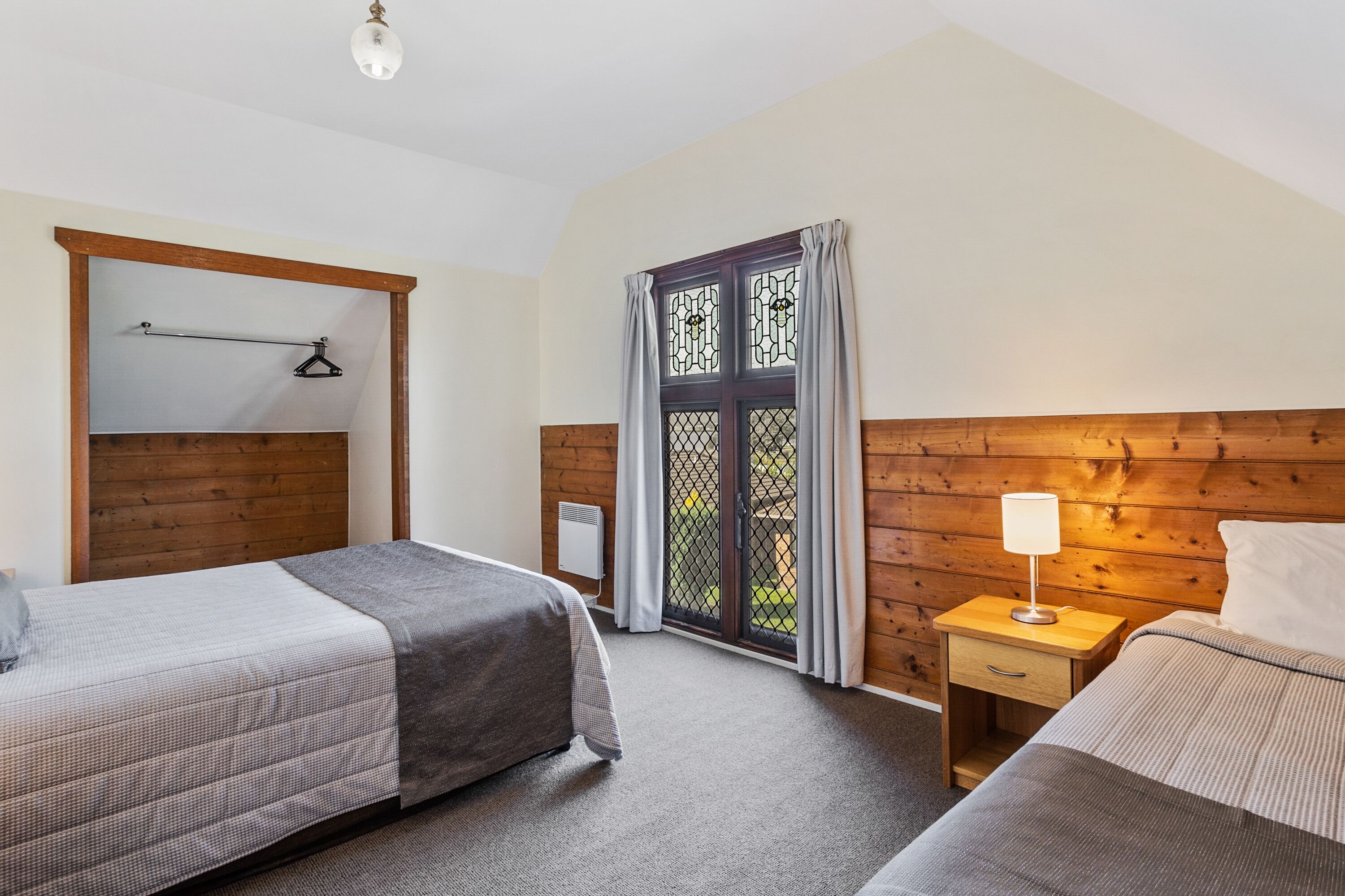 Ein Schlafzimmer mit zwei Betten, einem hölzernen Kopfteil, einem Nachttisch mit Lampe und einem Fenster mit Vorhängen.