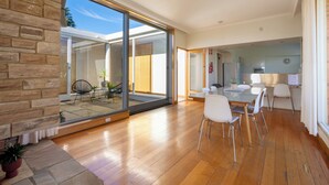 A spacious living area with a wooden floor, a glass sliding door leading to a balcony, and a dining area with white chairs and a table.