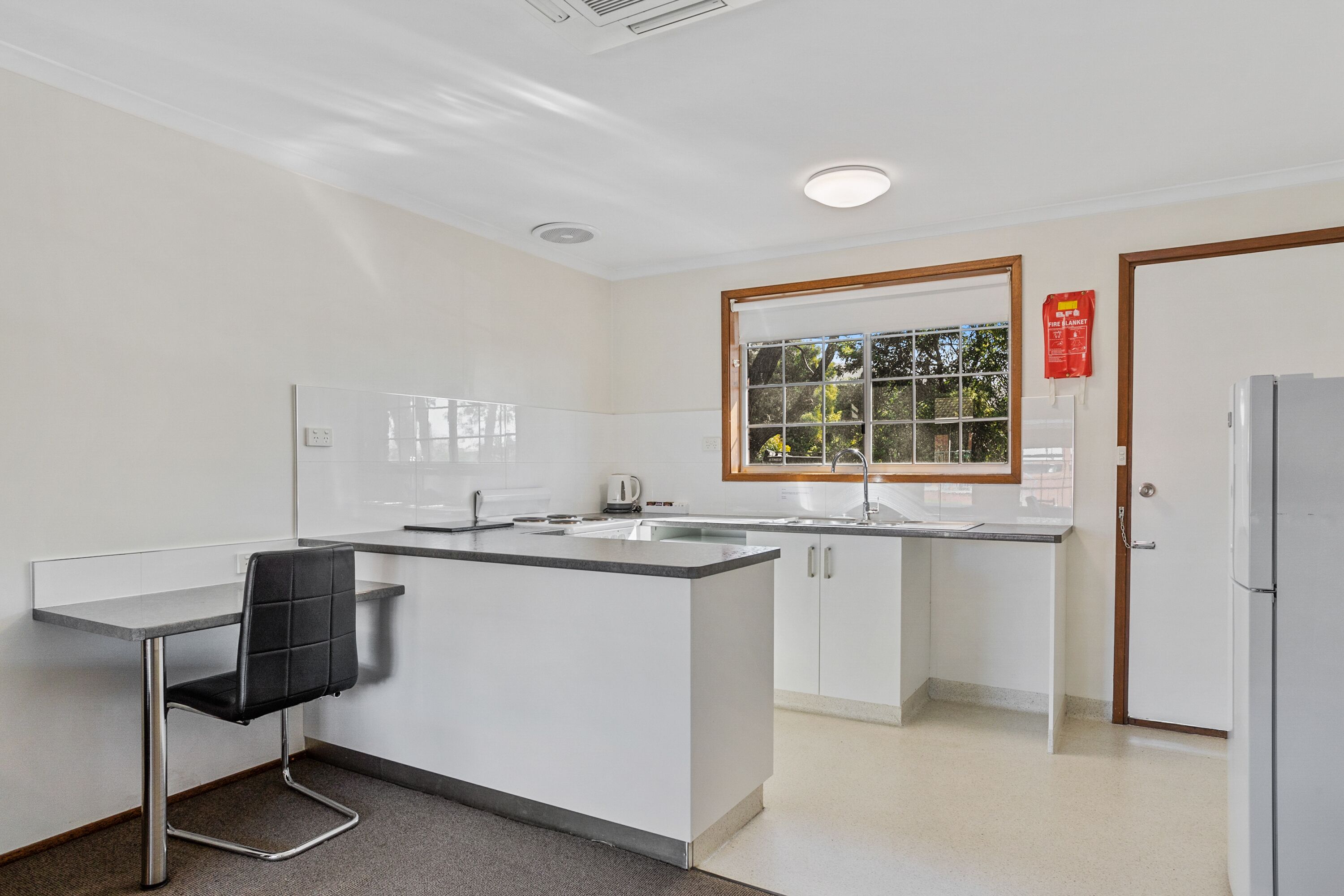 A modern kitchen with a black chair, white cabinets, a window, and a refrigerator.