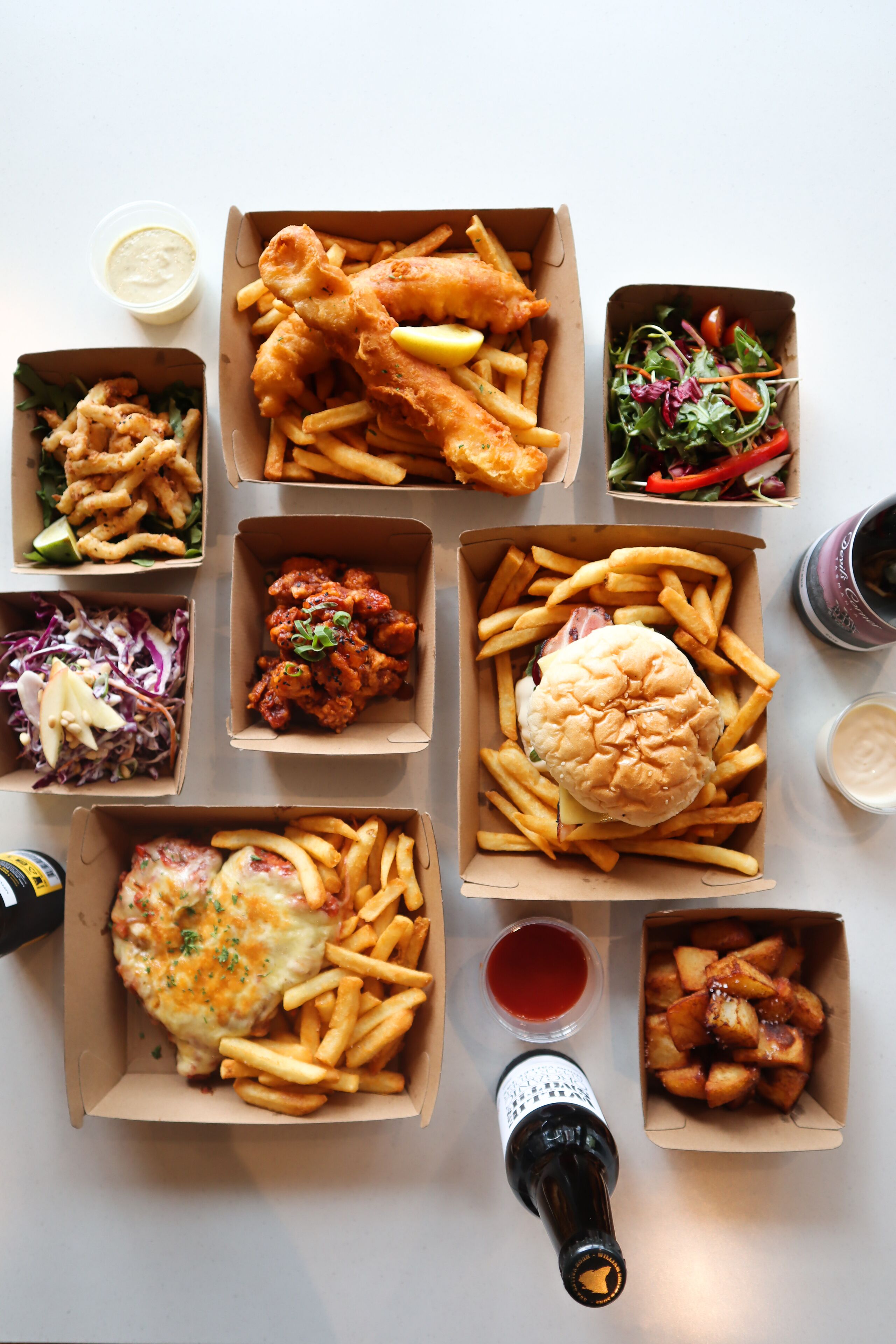 A variety of food items in cardboard boxes, including fish and chips, burgers, and fries, along with a bottle of beer and a cup of sauce.