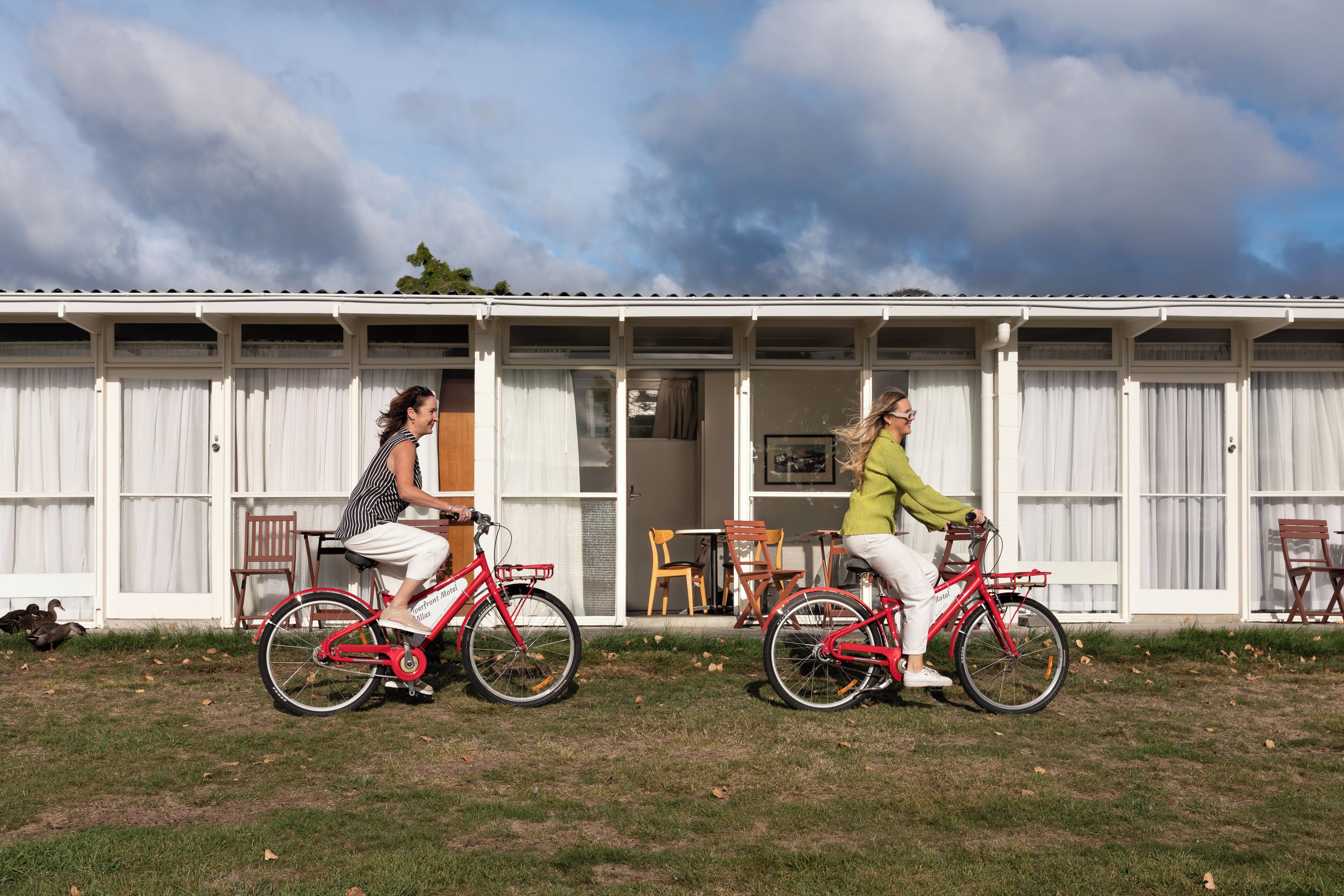 Two individuals riding red bicycles in front of a white building with a covered patio area.