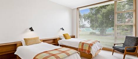 A hotel room with two beds, a chair, and large windows with blinds.
