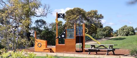Un parque infantil con una estructura de juegos temática de piratas, que incluye un tobogán y una zona de escalada, rodeado de árboles y una mesa de picnic.