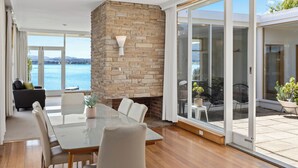 A modern dining area with a stone fireplace, a glass table, and chairs, overlooking a scenic view of the sea.