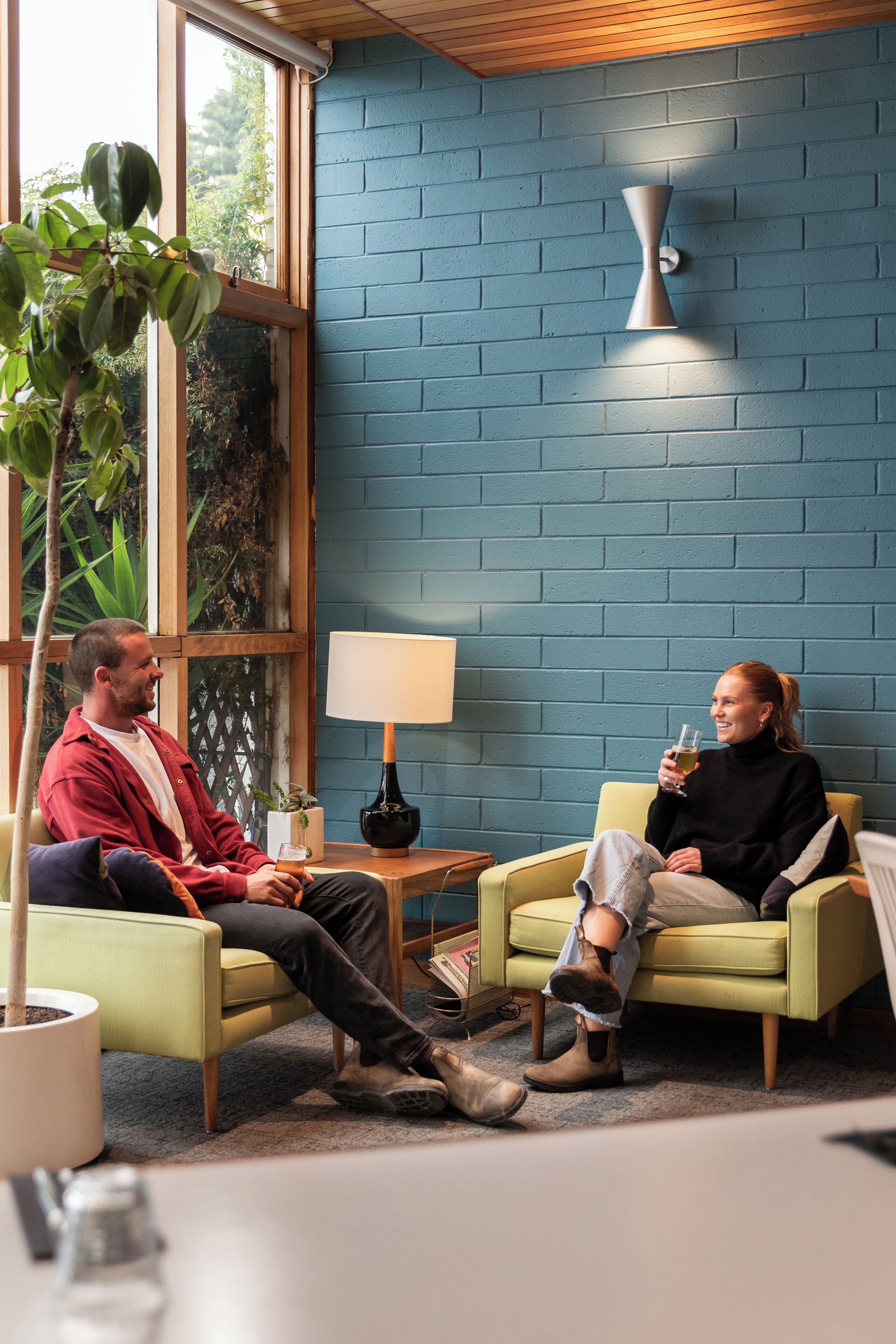 Dos personas sentadas en sofás verde claro en una habitación con pared de ladrillo azul, una planta en maceta y una lámpara de mesa.