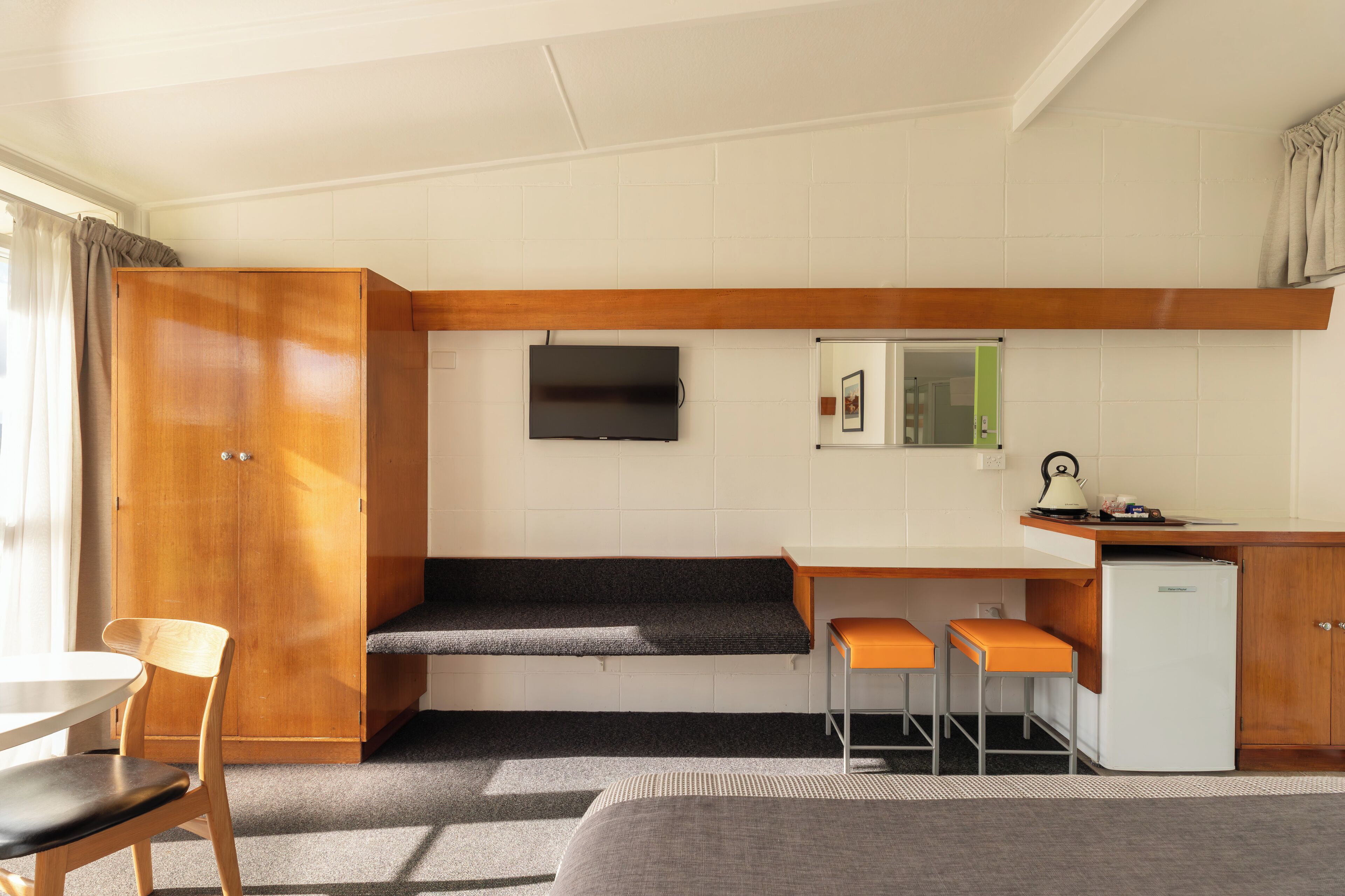 A compact kitchenette with a wooden cabinet, a small refrigerator, a TV mounted on the wall, and a bench with an orange seat.