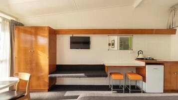 A compact kitchenette with a wooden cabinet, a small refrigerator, a TV mounted on the wall, and a bench with an orange seat.