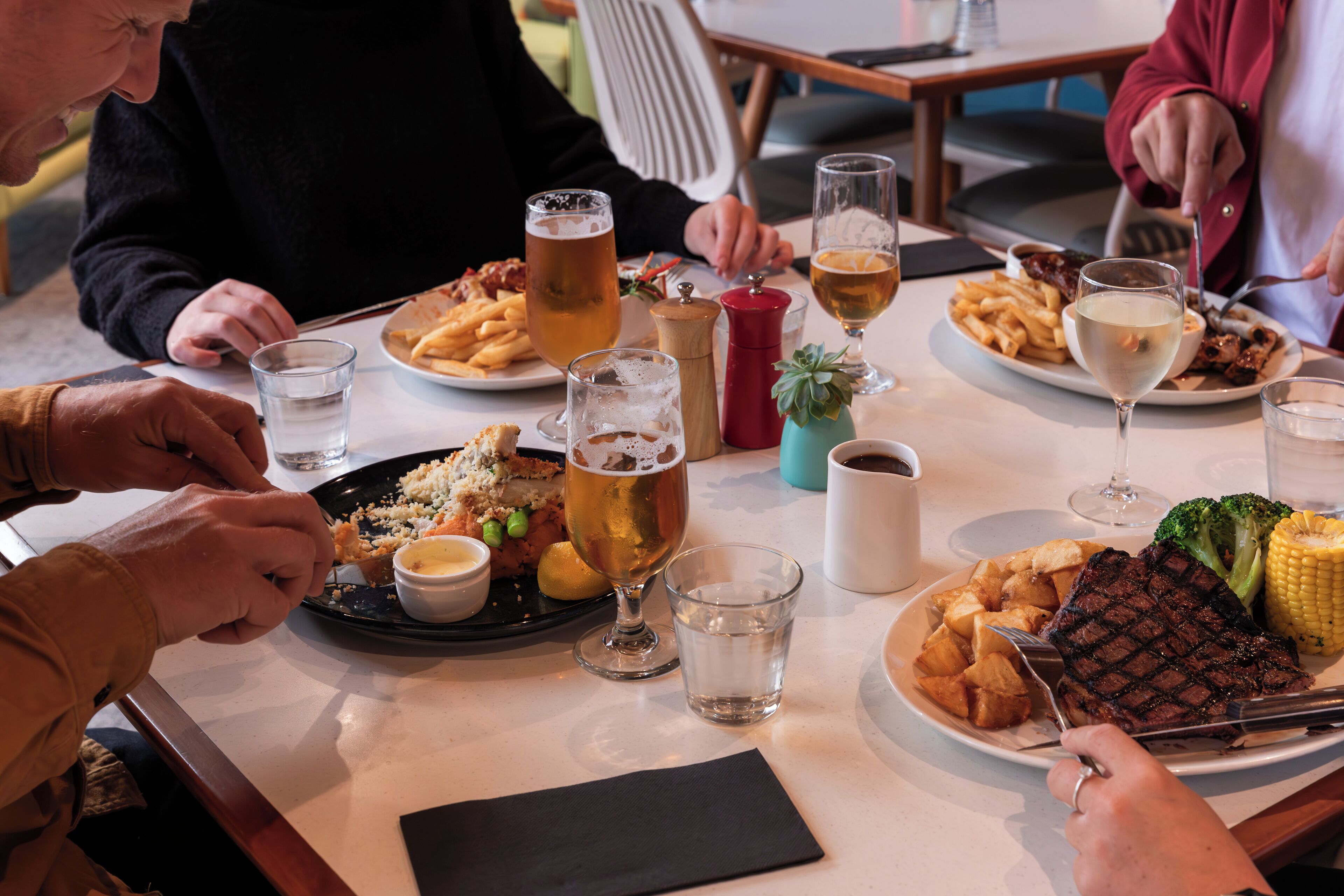 A dining table with plates of food, glasses of beer, and a steak.