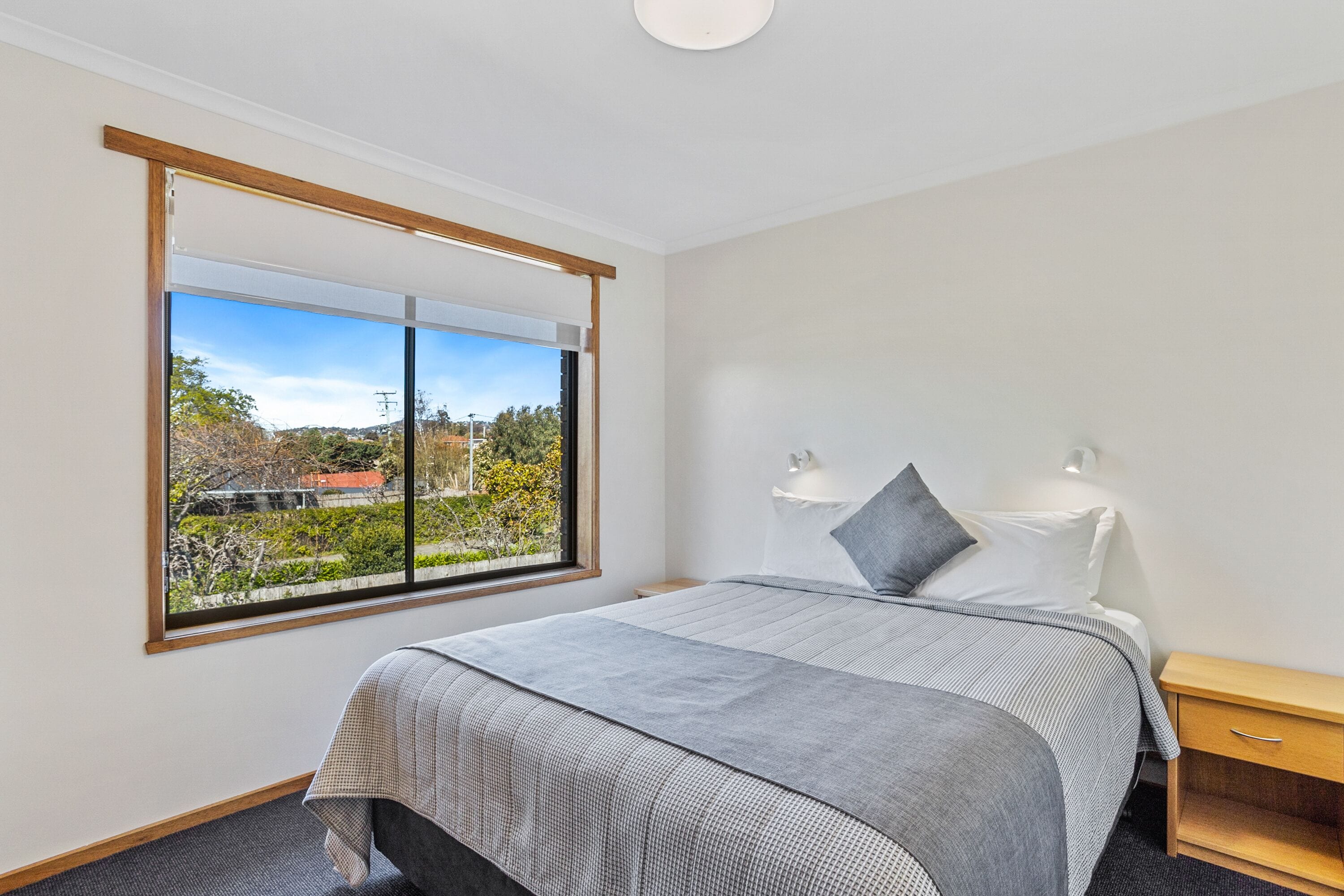 A modern bedroom with a large window, a bed with grey bedding, a bedside table with lamps, and a view of a park and buildings.
