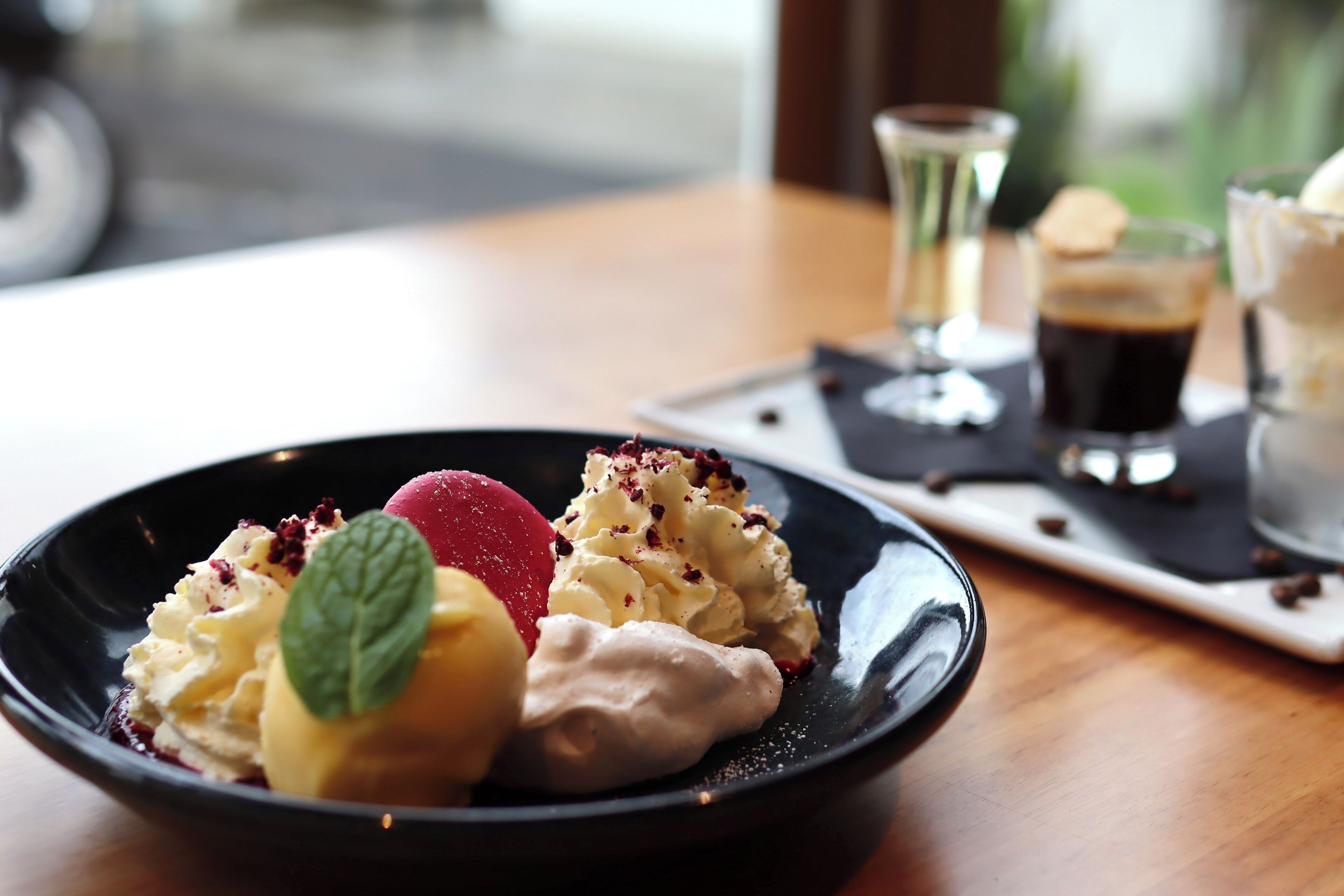 Une assiette de dessert avec différentes sortes de glaces et une feuille de menthe en guise de décoration, accompagnée de tasses à café et d’un petit verre sur une table en bois.