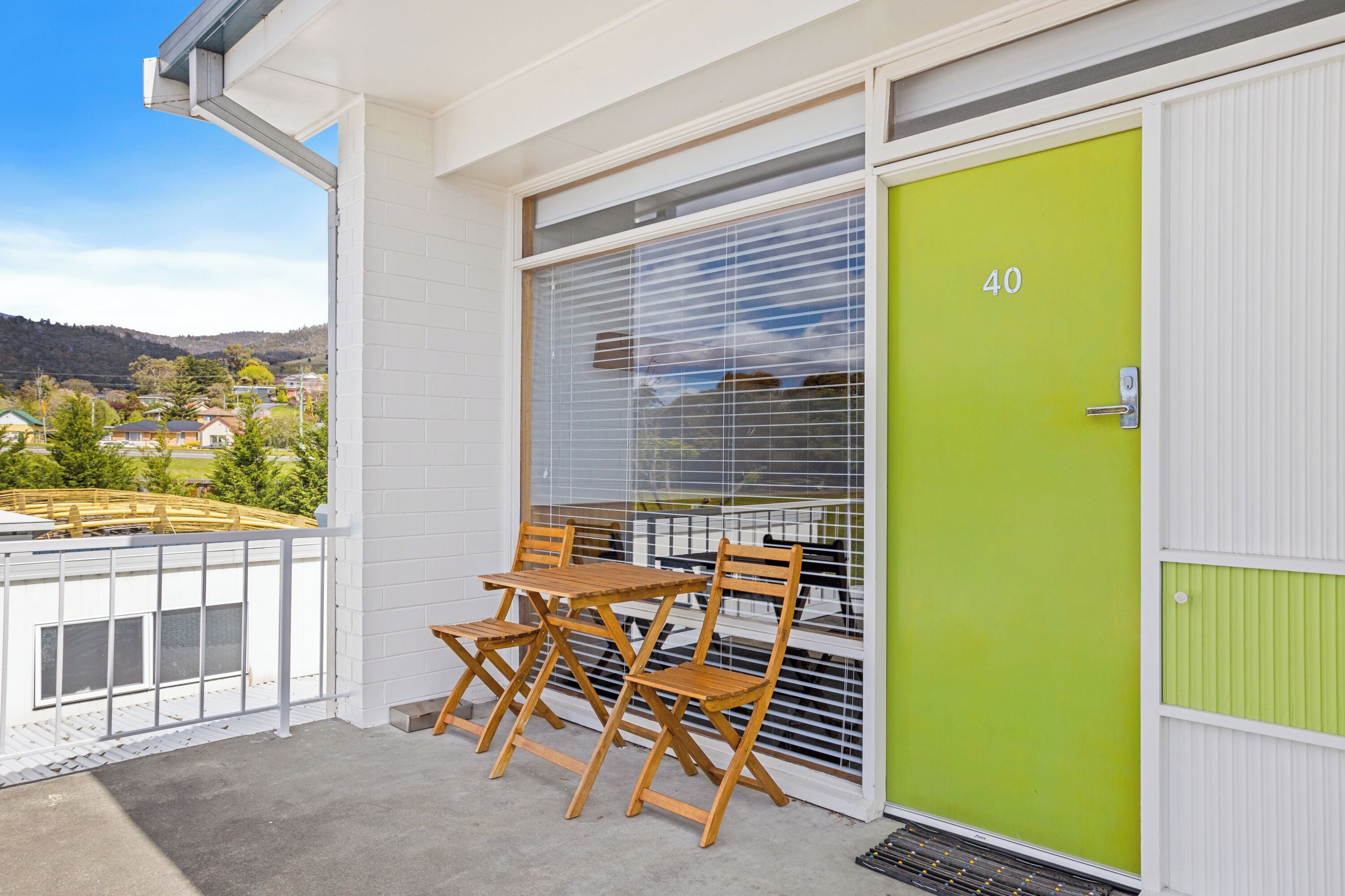 A balcony with a table and chairs, a green door with the number 40, and a view of houses and hills in the background.