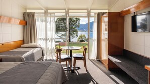 A hotel room with two beds, a small table with chairs, a TV, and a large window offering a view of a lake and mountains.