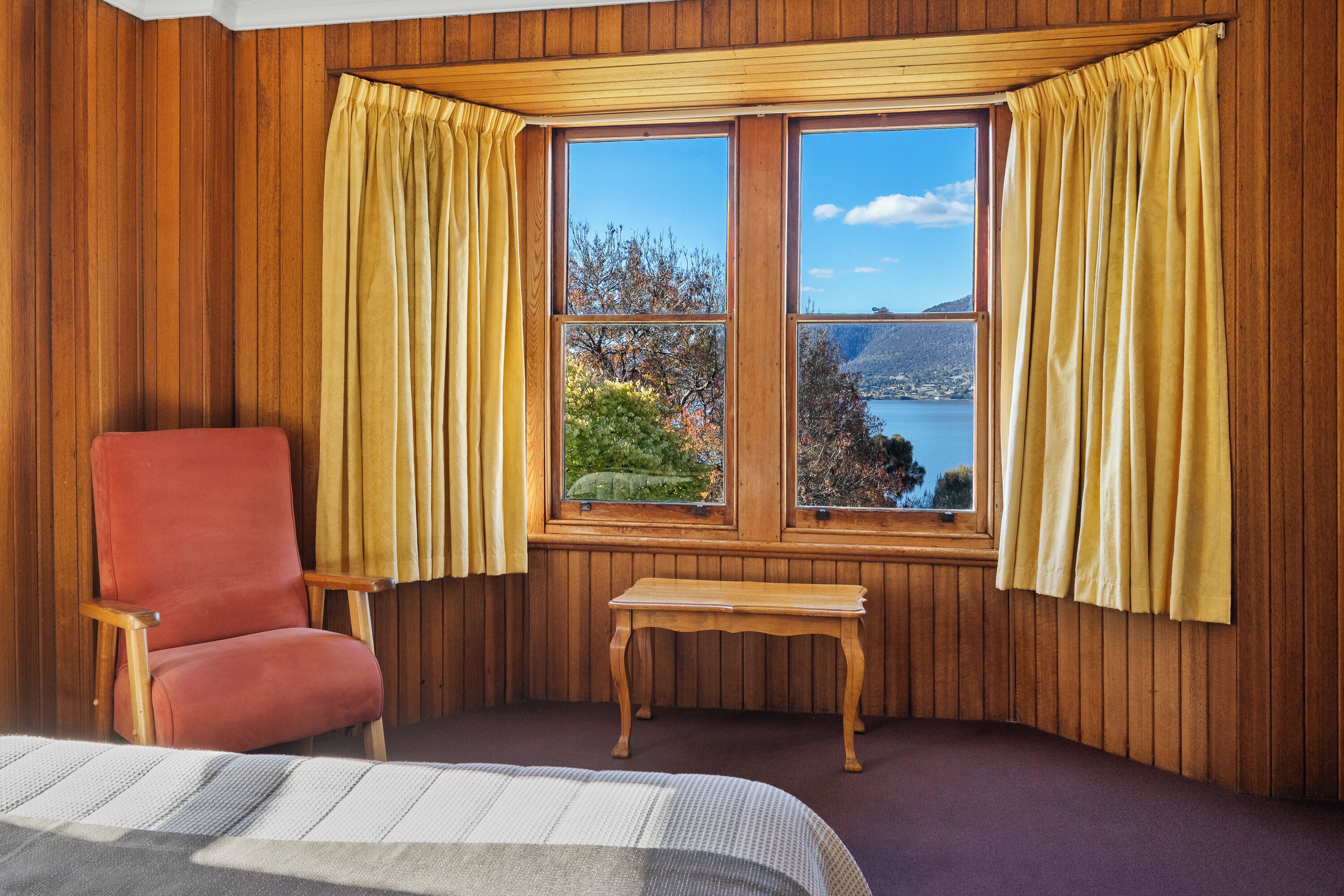 Ein Zimmer mit Holzwandverkleidung, einem roten Sessel, einem kleinen Holztisch und einem Fenster mit gelben Vorhängen.