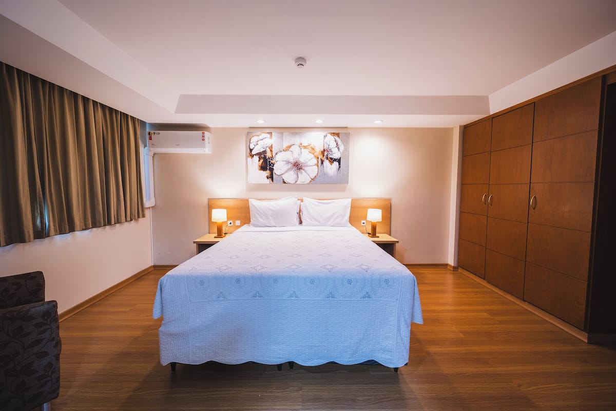 A bedroom with a large bed, two bedside tables with lamps, a wardrobe, and a wall-mounted air conditioner.