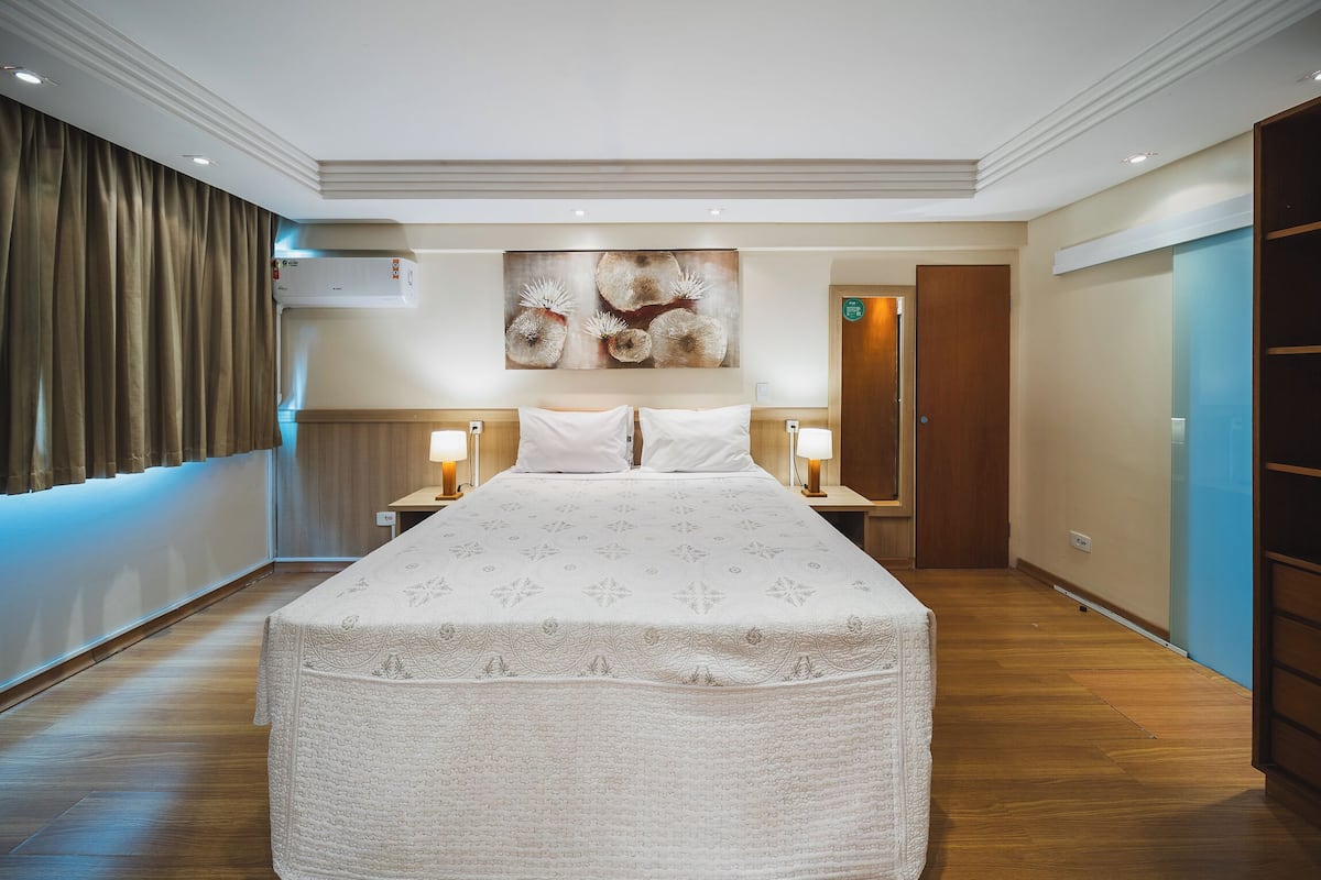 A hotel room with a large bed, two bedside tables with lamps, a wall-mounted air conditioner, and a large abstract painting above the bed.