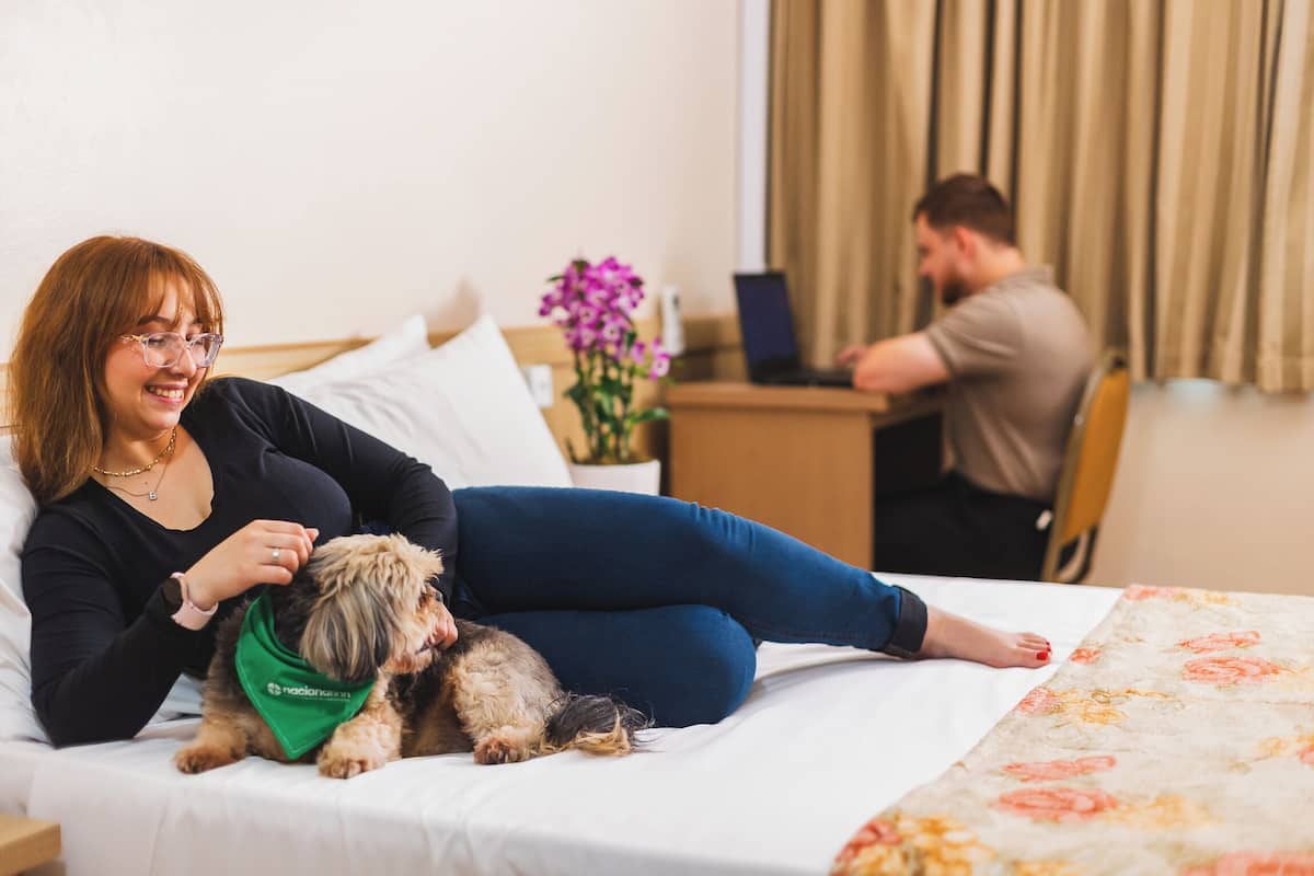 A hotel room with a bed, a desk with a laptop, and a person sitting at the desk.