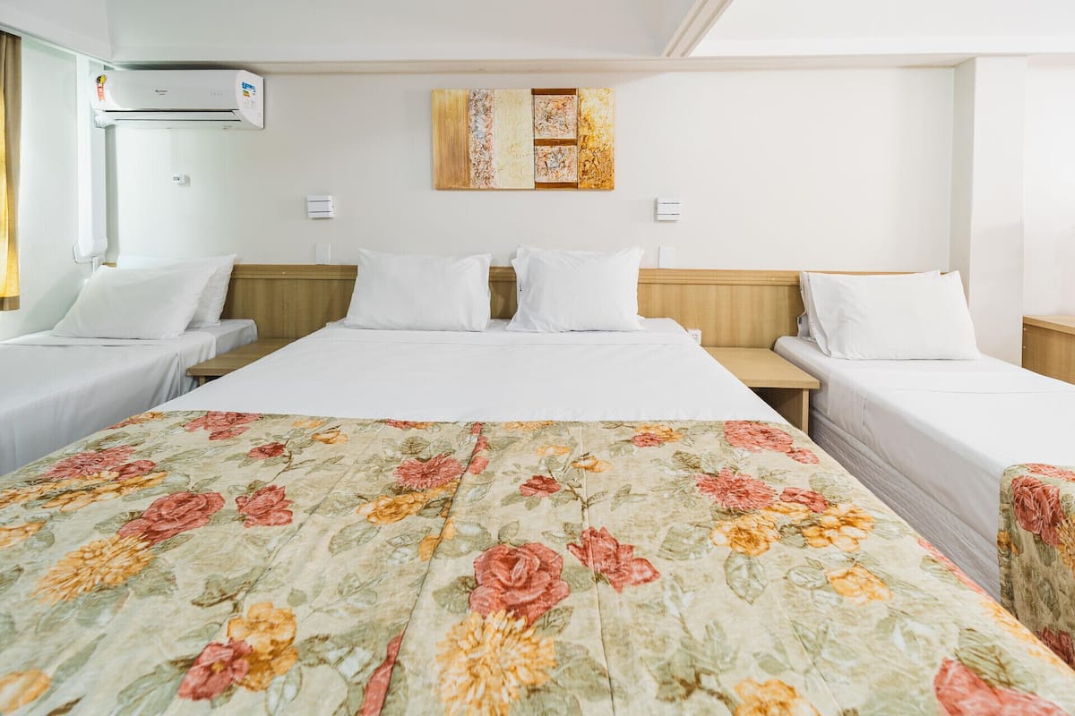 A hotel room with a large bed, two single beds, a wall-mounted air conditioner, and a painting.
