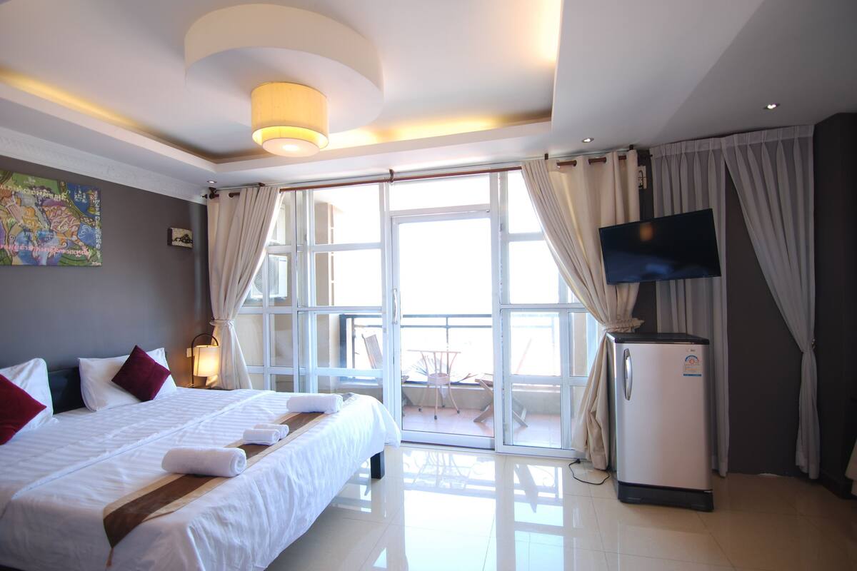 A hotel room with a bed, a TV, a refrigerator, and a balcony with a table and chairs.