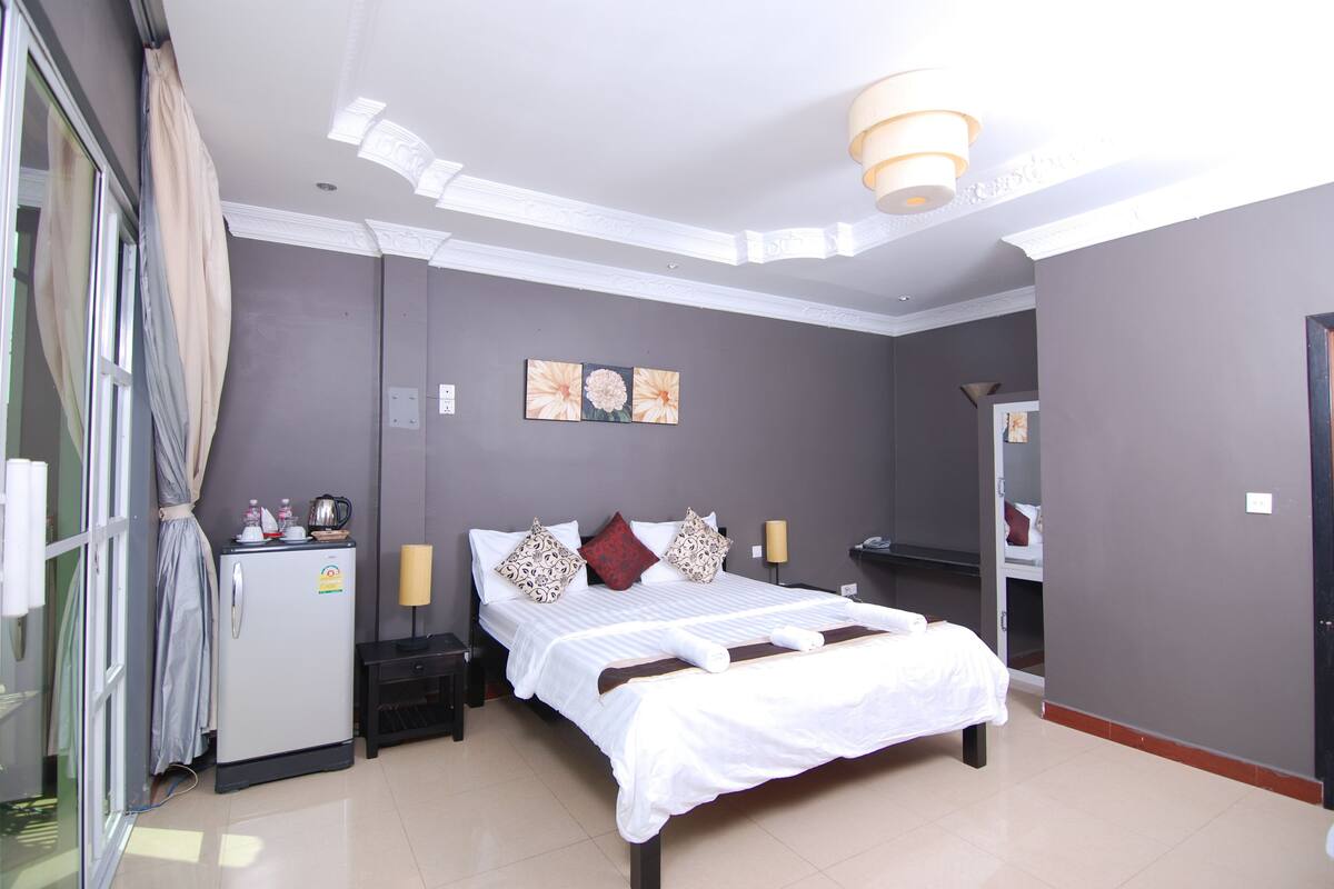 A hotel room with a bed, a small refrigerator, a bedside table, and a mirror.