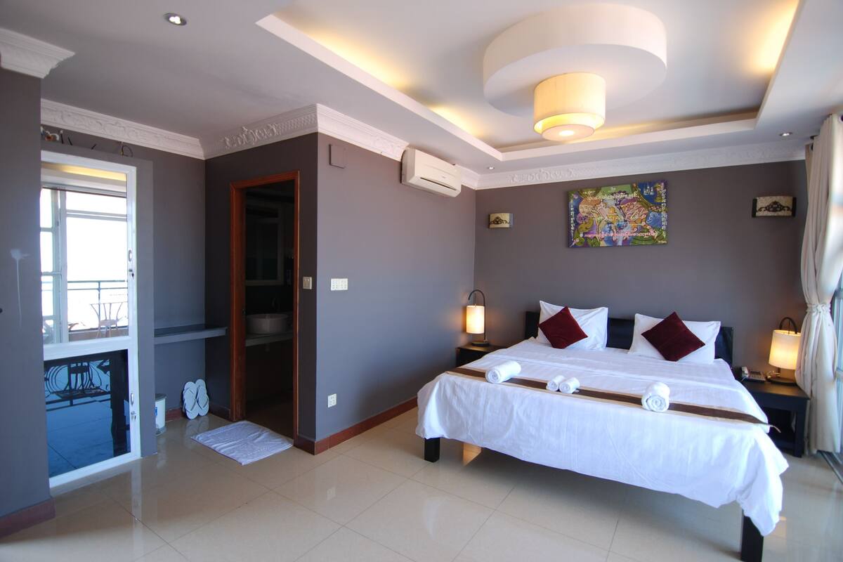 A hotel room with a large bed, two bedside tables with lamps, a ceiling light, an air conditioning unit, a painting on the wall, and a door leading to another room.