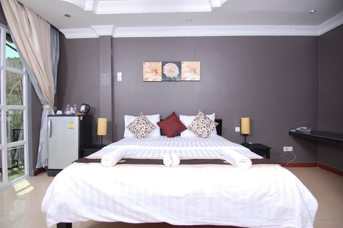 A hotel room with a large bed, a small refrigerator, a desk with a lamp, and a window with curtains.