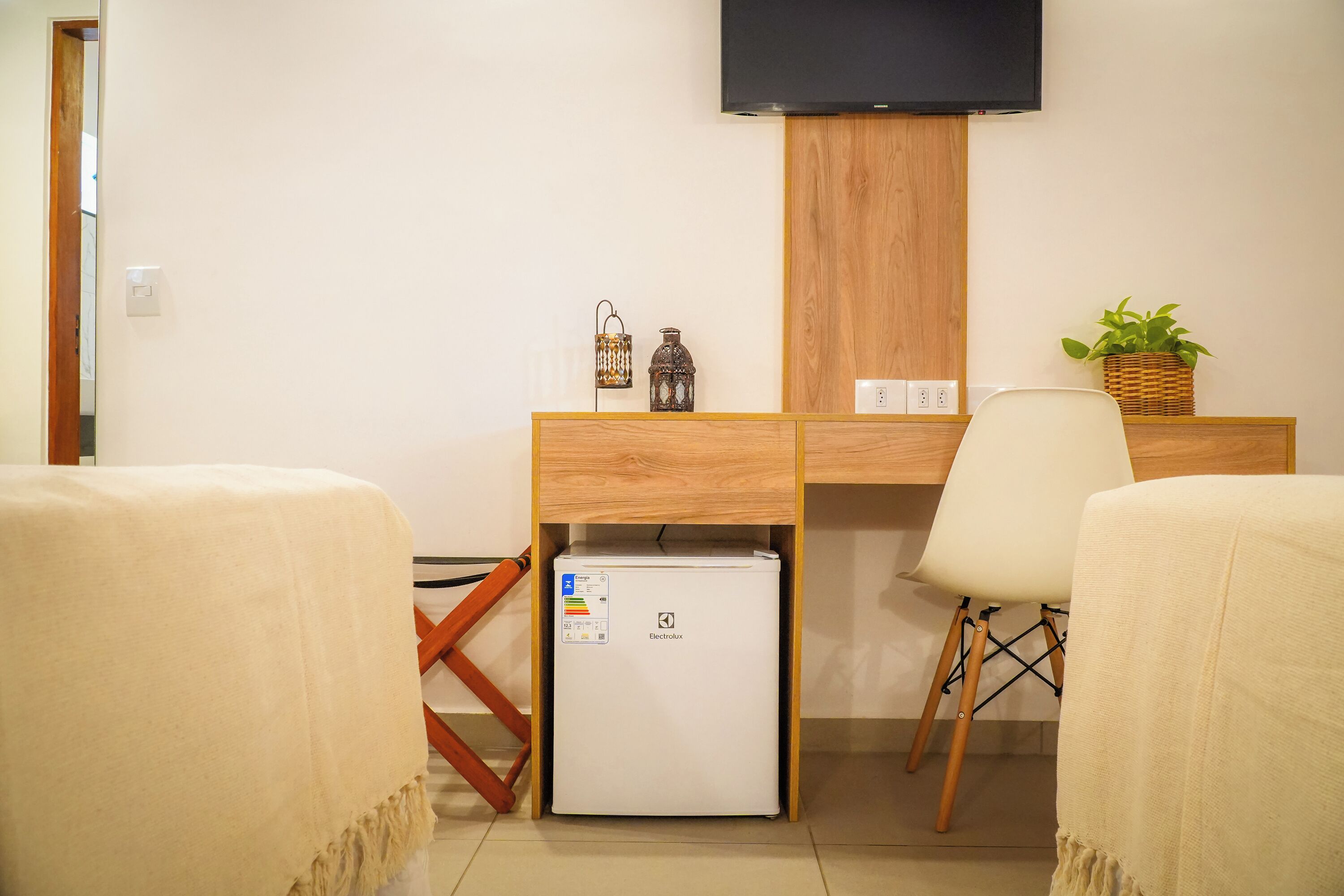 Habitación de hotel con escritorio de madera, refrigerador pequeño, televisor montado en la pared y una planta en maceta.