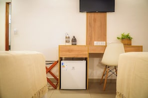 Habitación de hotel con escritorio de madera, refrigerador pequeño, televisor montado en la pared y una planta en maceta.