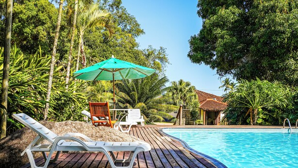 Een houten terras met lounge stoelen en een parasol, een zwembad en weelderige tropische beplanting.