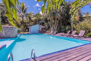 Una piscina con muelle de madera, sillas de descanso y una pequeña estructura, rodeada de vegetación tropical.