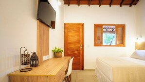 Une chambre avec un bureau en bois, un téléviseur fixé au mur, un lit recouvert d’une courtepointe blanche et une fenêtre avec des volets.