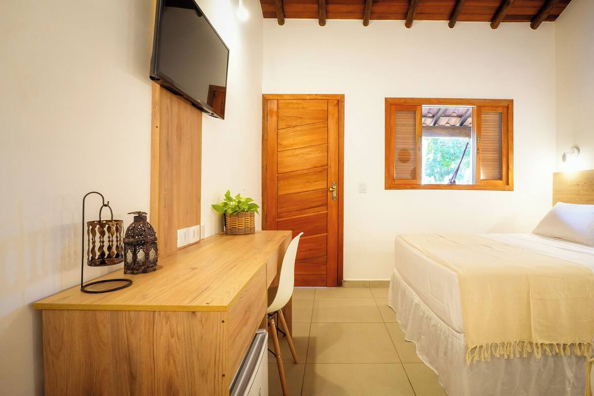 Ein Schlafzimmer mit einem Holztisch, einem an der Wand montierten Fernseher, einem Bett mit weißer Bettdecke und einem Fenster mit Fensterläden.