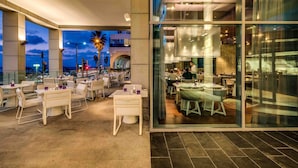 Un comedor interior con mesas y sillas, ventanas amplias y vistas a la ciudad al atardecer.