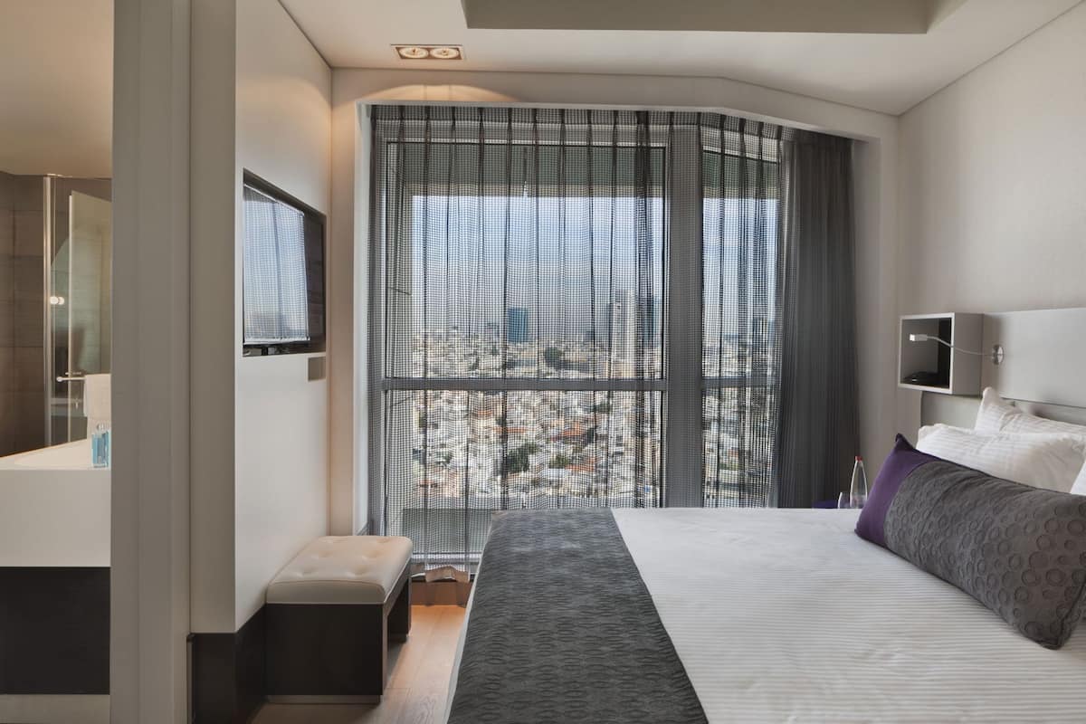 A modern hotel room with a large bed, a small bedside table, a built-in TV, and a large window with a view.