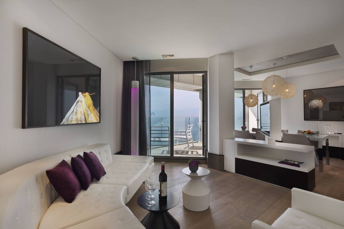 A modern living room with a white sofa, a dining area with a white table, and a view of the ocean through large glass doors.