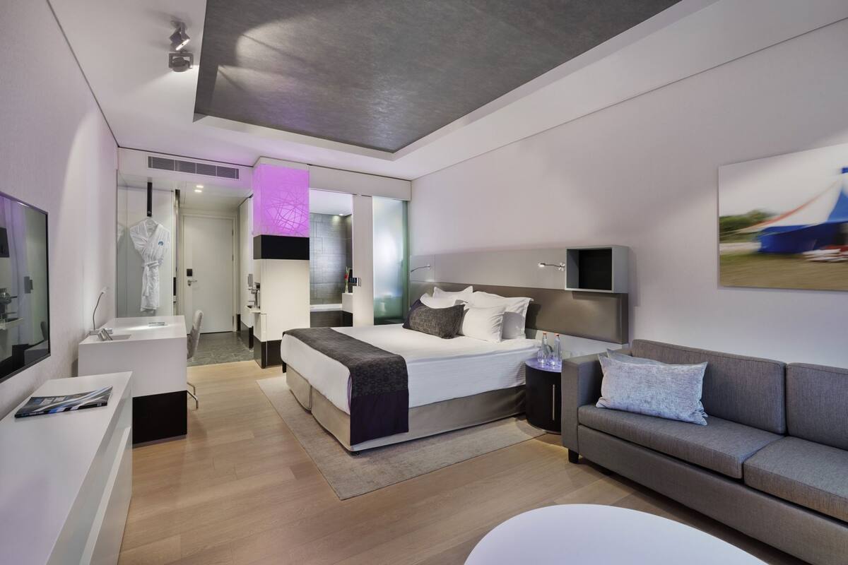 A modern hotel room with a bed, a sofa, a desk, and a bathroom visible through a glass door.