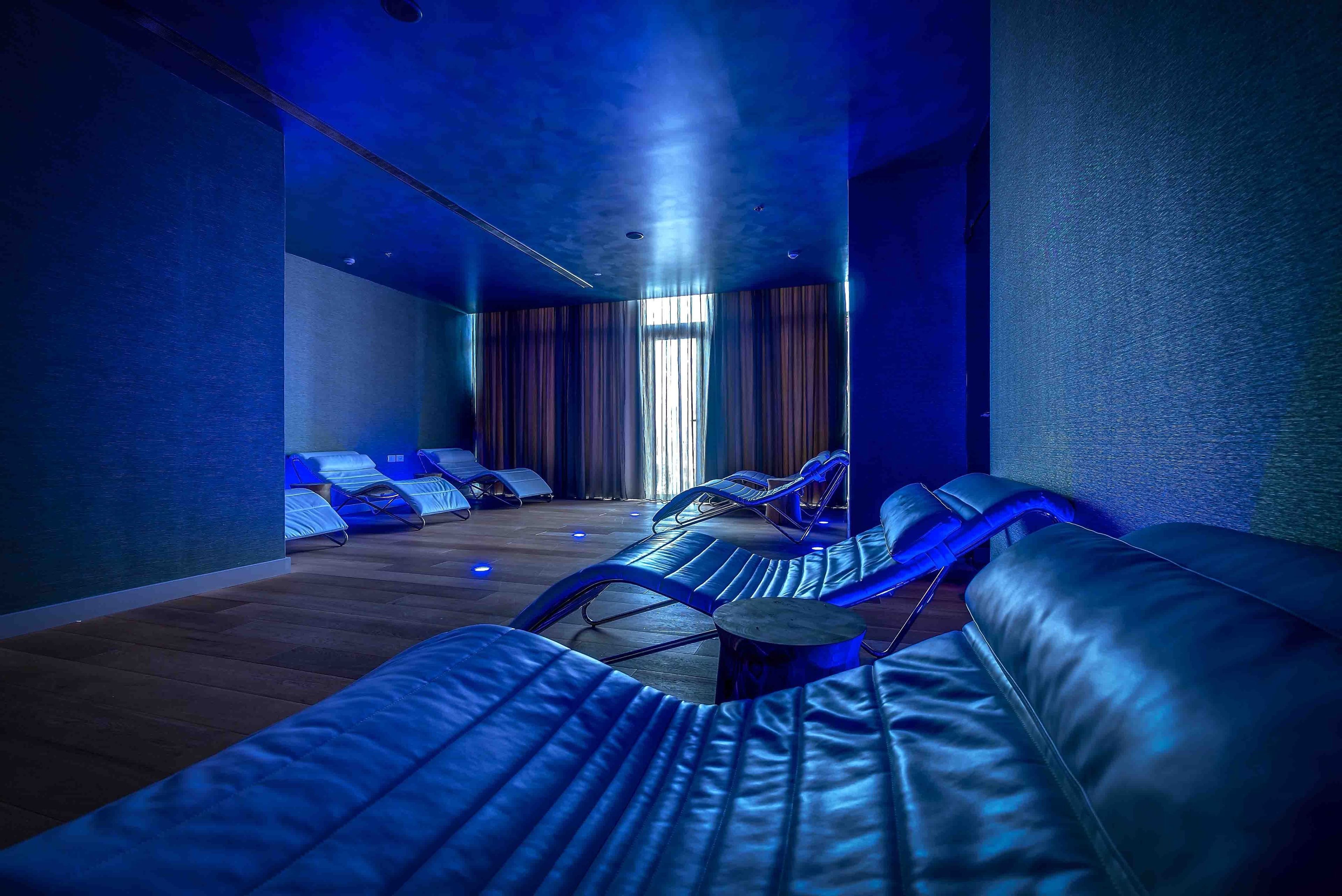 A room with blue ambient lighting, leather reclining chairs, a wooden floor, and a large window with curtains.