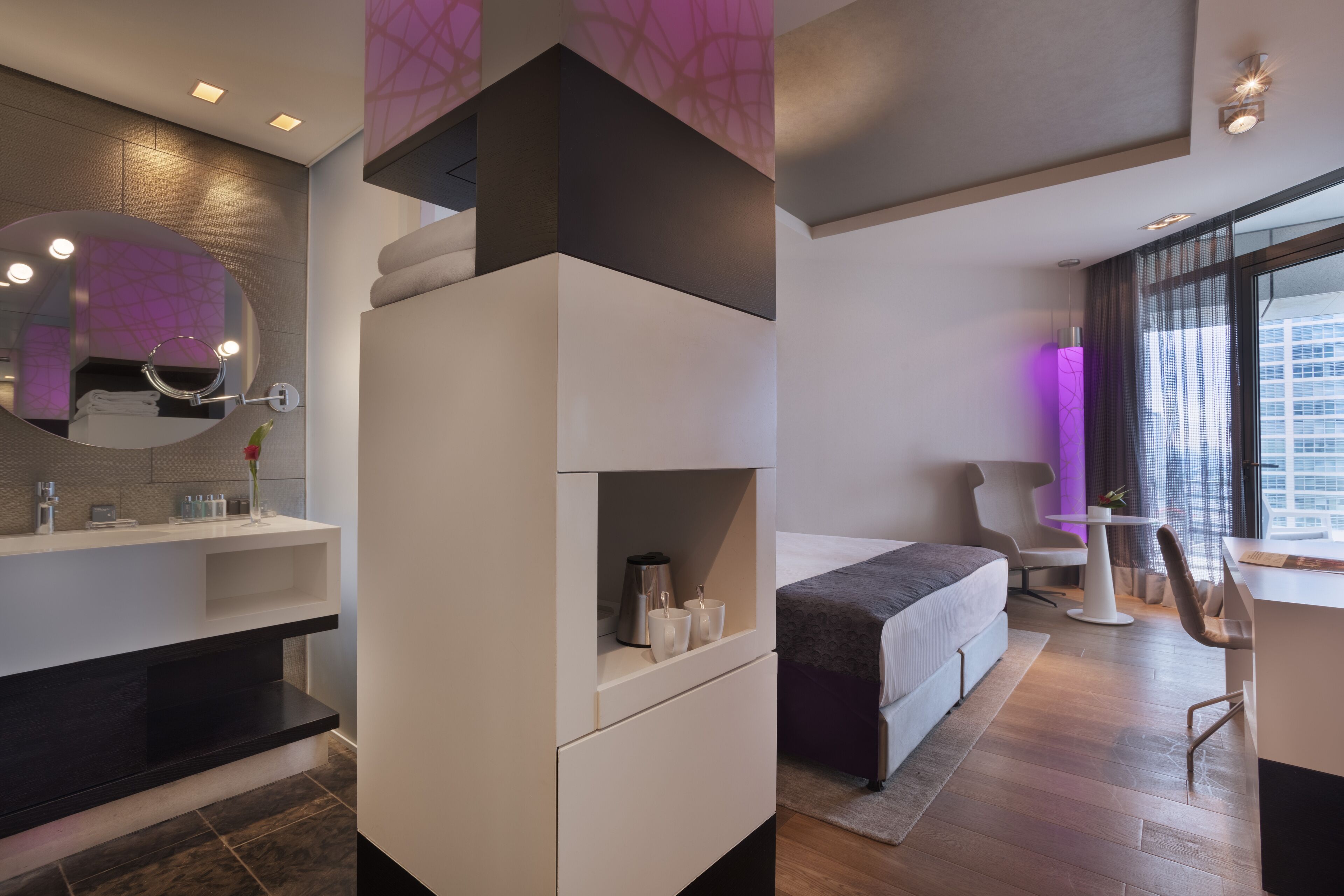 Habitación de hotel moderna con un espejo grande, tocador con artículos de aseo, cama con cabecera gris, una mesita y vistas a la ciudad a través de amplios ventanales.