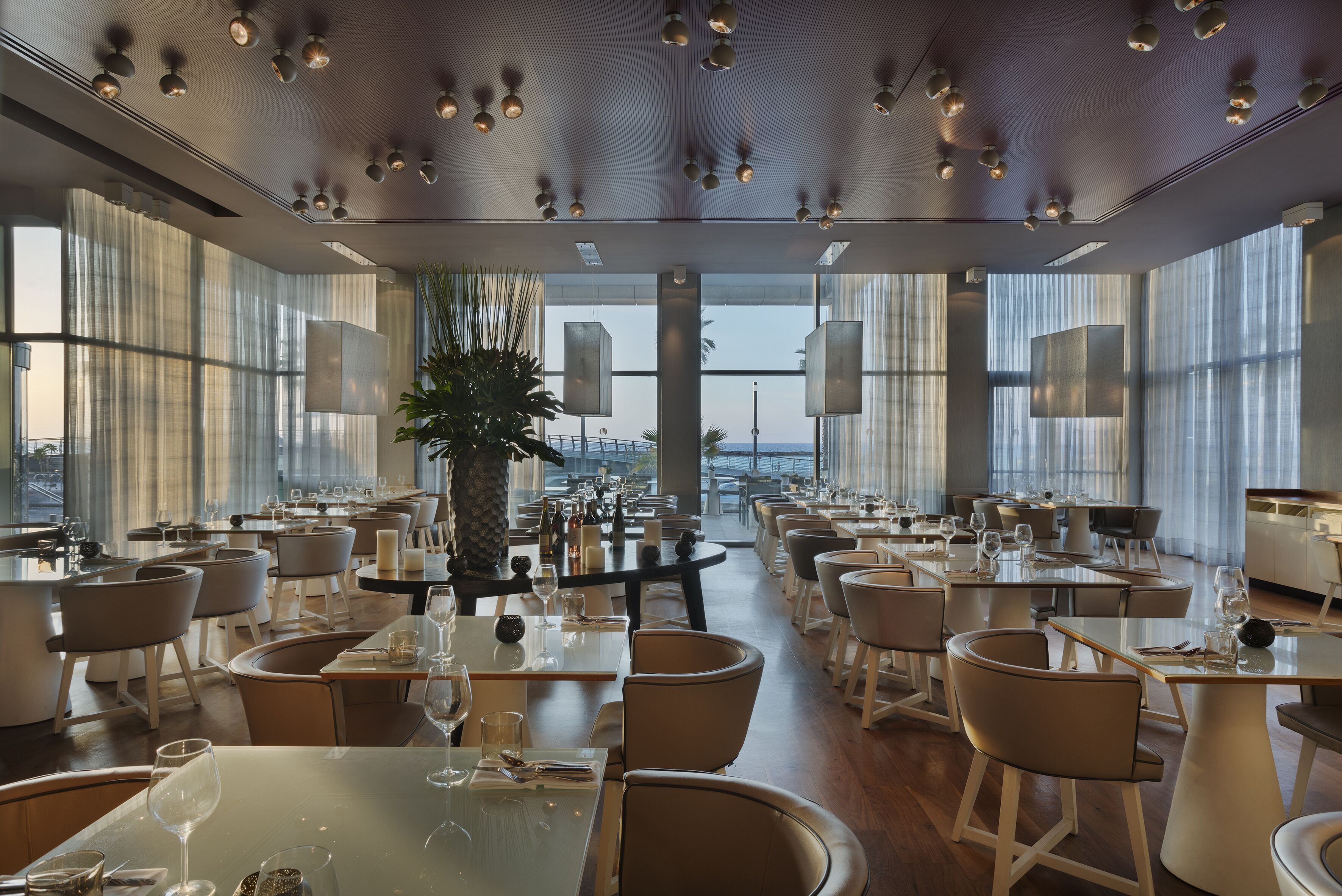 Un comedor amplio con varias mesas y sillas, ventanas grandes y vista panorámica de la ciudad.