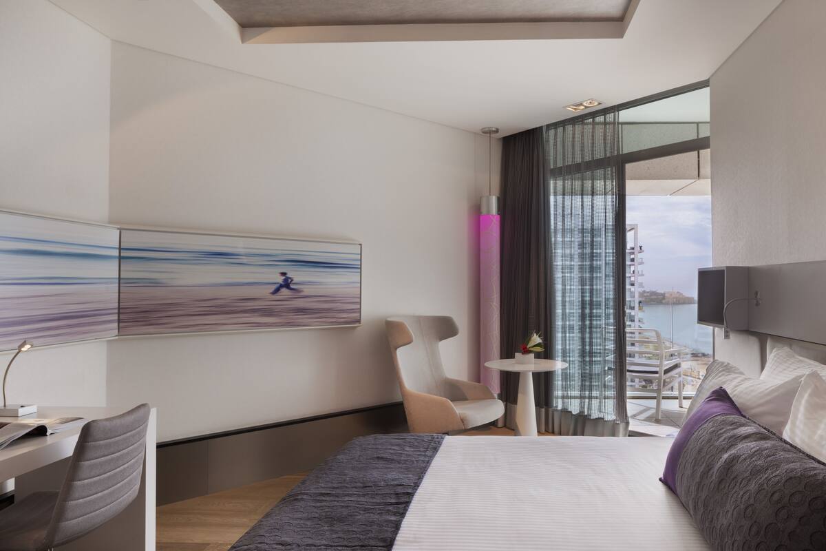 A modern hotel room with a large bed, a desk, a chair, and a small table. There is a painting on the wall and a view of the city through the window.
